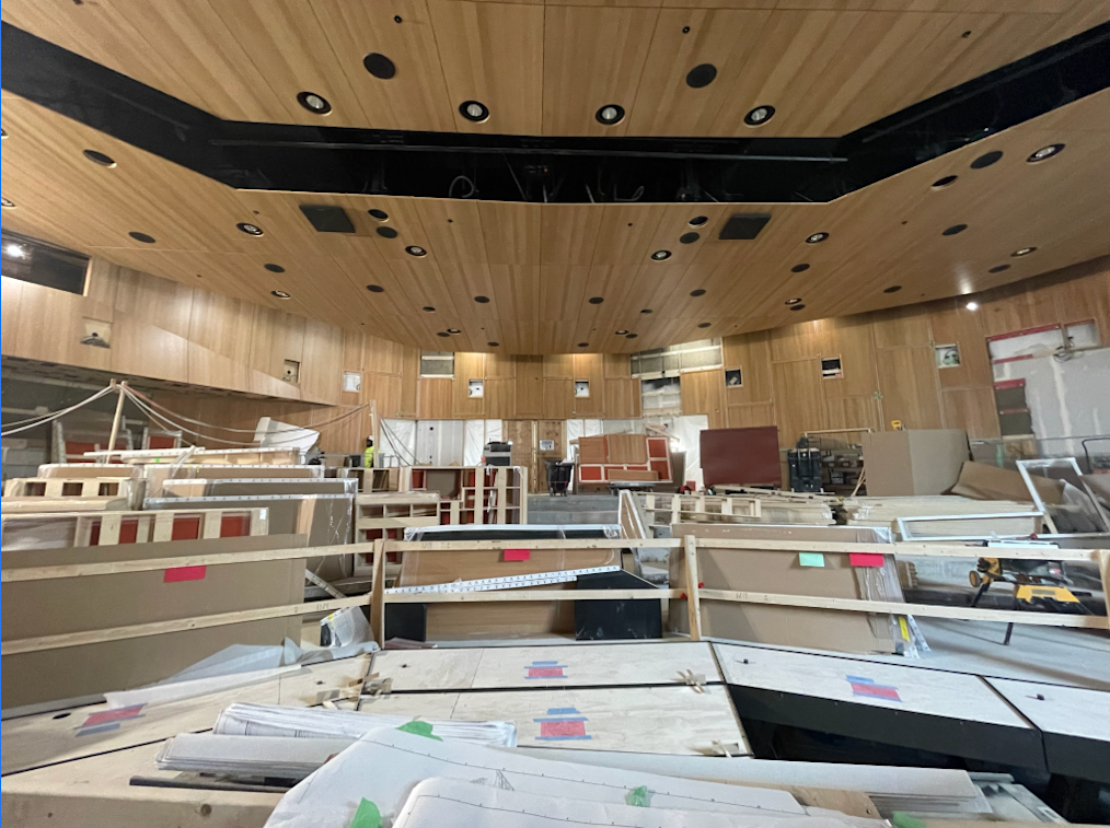 An image of construction of the Elie Wiesel Auditorium. The top and the back wall of the Auditorium is covered in brown wood paneling with light fixtures. In the center of the image are several beams and pieces of wood. These pieces will become the stage and seats in the Auditorium.