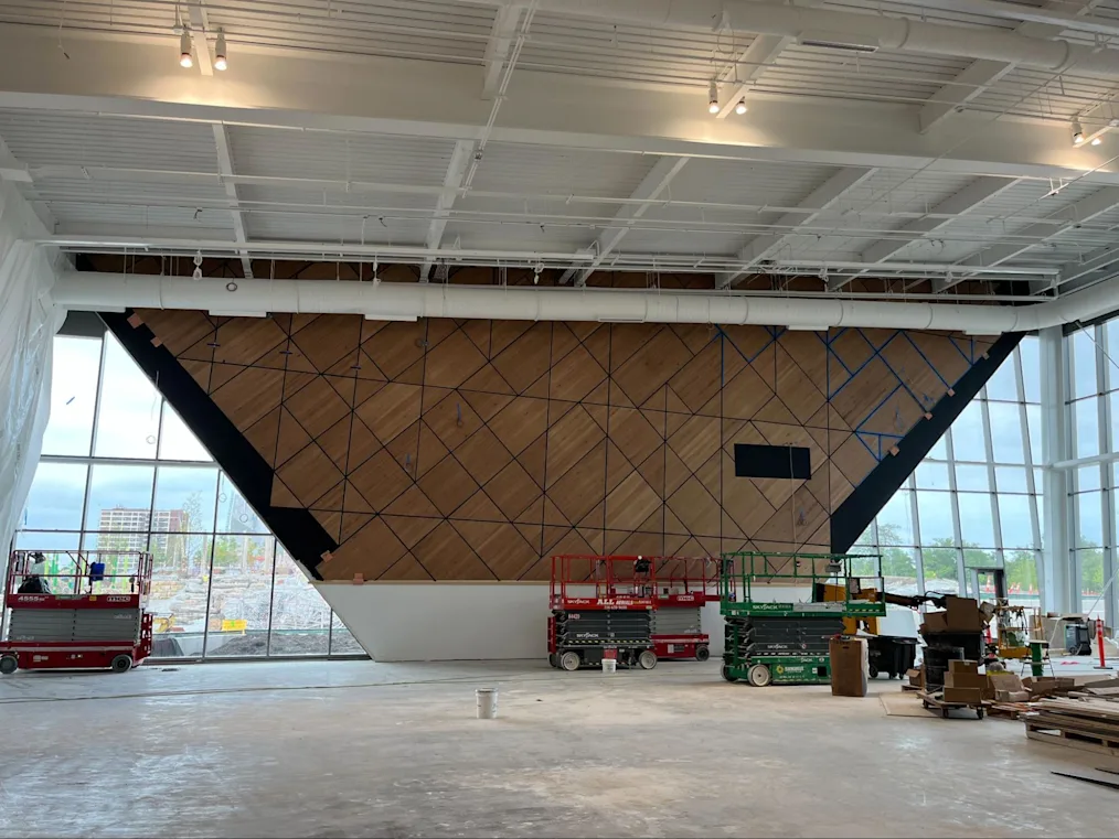 An image of construction being done on Main Court, the Obama Presidential Center's basketball court, at Home Court. The wood panels are on one wall and are brown with blue borders. Each panel of wood is a triangular or diamond shape. Next to the wood panel wall is a glass window. In front of the off the wall are two construction workers on a red crane installing the panels.