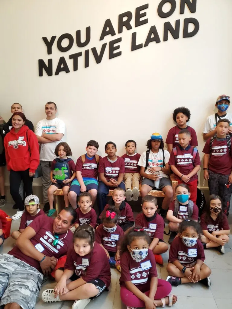 A group of young boys and girls of a range of skin tones and ages sit with their legs crossed in front of a sign at the Chicago Field Museum. The sign reads, “you are on native land.”