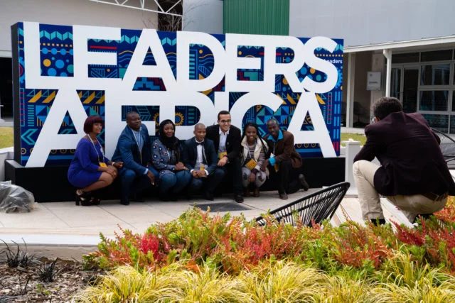 

A group of men and women ranging from medium to deep skin tone kneels outdoors in front of a blue sign with white letters that reads "Leader Africa" as someone takes their picture 