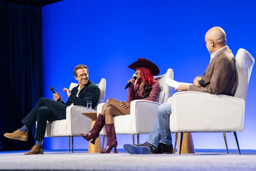 Ryan Reynolds sits on a stage with Reyna Roberts and Ayad Akhtar at the 2024 Democracy Forum. He is holding a microphone. 