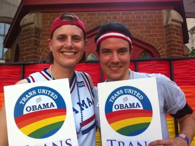 The image is a candid shot of Dylan Orr with a friend. Dylan has an olive complexion and short dark wavy hair. Dylan is wearing a red and white striped headband and a gray t-shirt. In the picture, Dylan and his friend are holding up signs that say "Trans United for Obama." In the sign is the Obama for America logo with a Pride rainbow in the center.