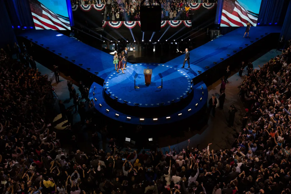 The first family walks onto a stage with bright blue walkways in front of a large crowd.
