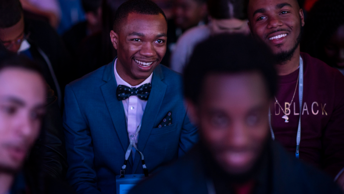 A black man wearing a suit with bowtie and pocket square smiles broadly. Next to him is another Black man with a maroon t-shirt, also smiling but looking to the left. Other people are seated around them.