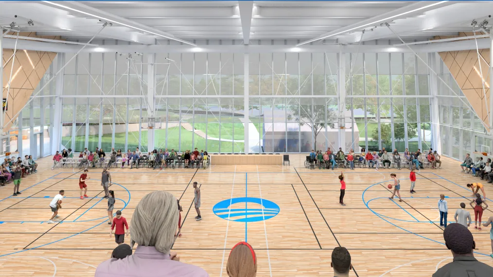 The image is a rendering of our Home Court gymnasium. The gymnasium has a wooden floor with basketball court markings and the Obama Foundation logo in blue at center court. The walls of the gymnasium are paneled in wood and light colored metal trusses span the ceiling. The wall furthest from the photo is made of rectangular glass windows. Through the windows you can see Jackson Park. In the gymnasium are people playing on the basketball court, people sitting in seats on the court, and people sitting in bleachers. 
