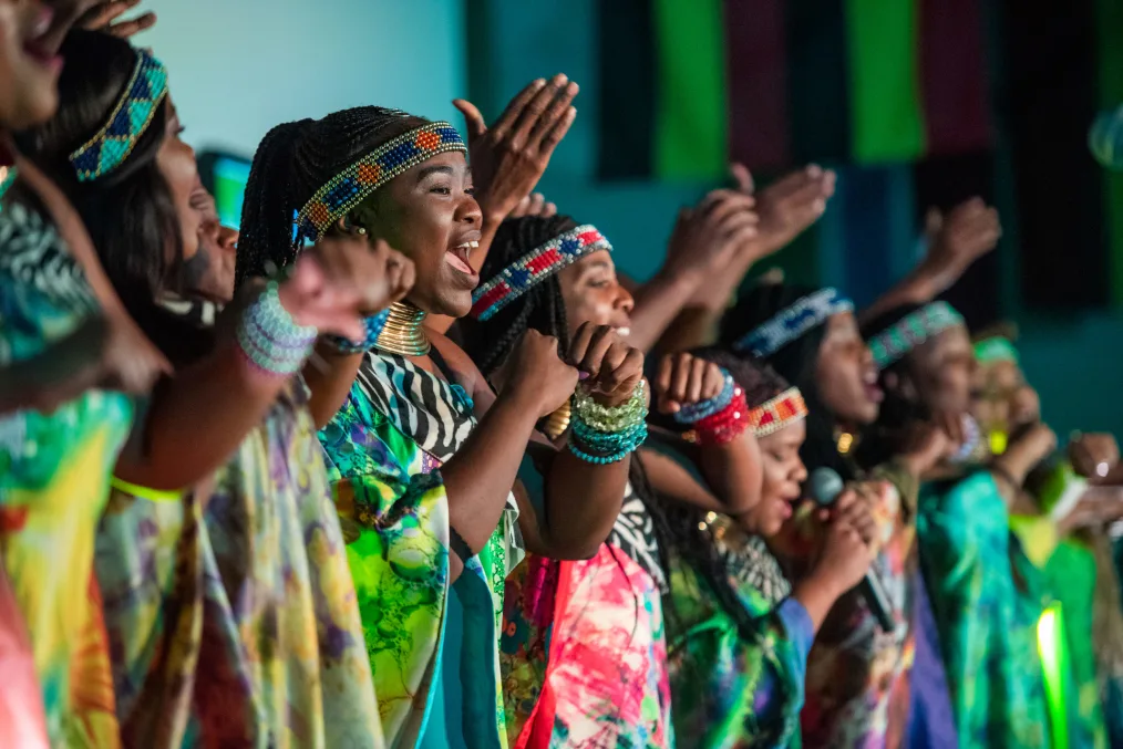 A group of women in colorful robes sing and raise their hands in praise.