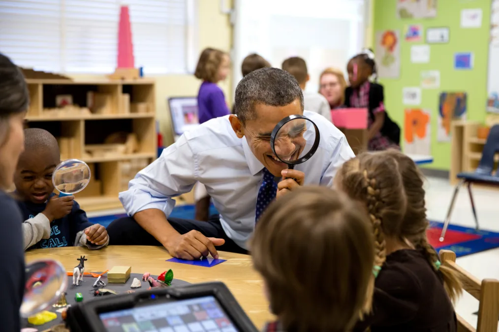 President Barack Obama, wearing a light blue button-up shirt and a blue tie with white specks, holds a black magnifying glass and looking toward a young girl with a light skin tone. There is a young boy sitting on the left hand side of President Barack Obama with a deep skin tone and a blue shirt with gray sleeves holds a blue and clear magnifying glass looking toward toys that are sitting on the table in front of them. There are other young individuals of a variety of skin tones blurred out in the photo.