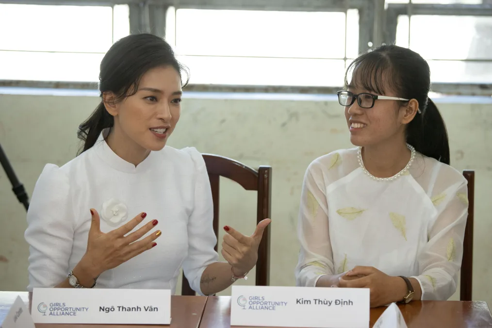 Two East-Asian woman, with long, black, tied back hair, have a conversation. They are wearing matching white dresses. The girl on the left eyeliner and her nails painted red with a golden nail on her ring finger and is talking at the moment. The girl on the right has glasses, bangs, and listens with a smile. They are seated a brown table. Their white name cards are on the desks infront of them with the Girls Opportunity Alliance logo in the corner. The girl on the left name is "Ngo Thanh Van" and on the right her name is "Kim Thuy Dinh."
