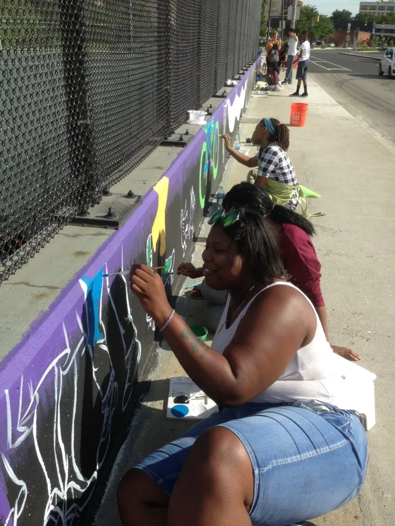Several people paint a low, cement wall in bright colors along a street.