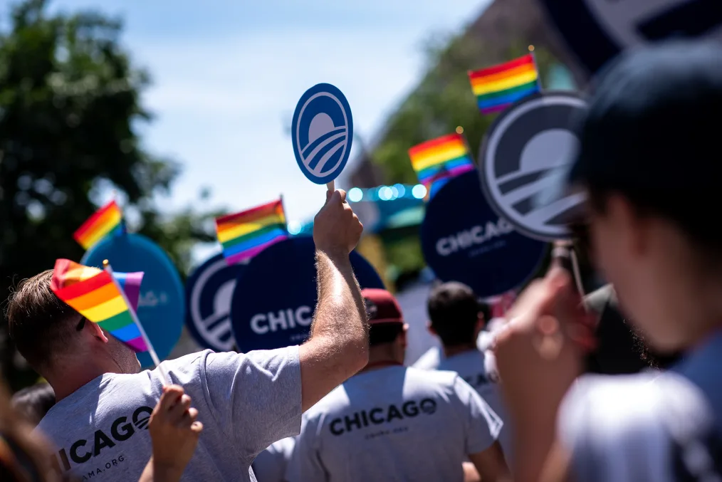 
An image of various people wearing gray shirts with "Chicago" on the back hold pride flags and Obama Foundation logo signs