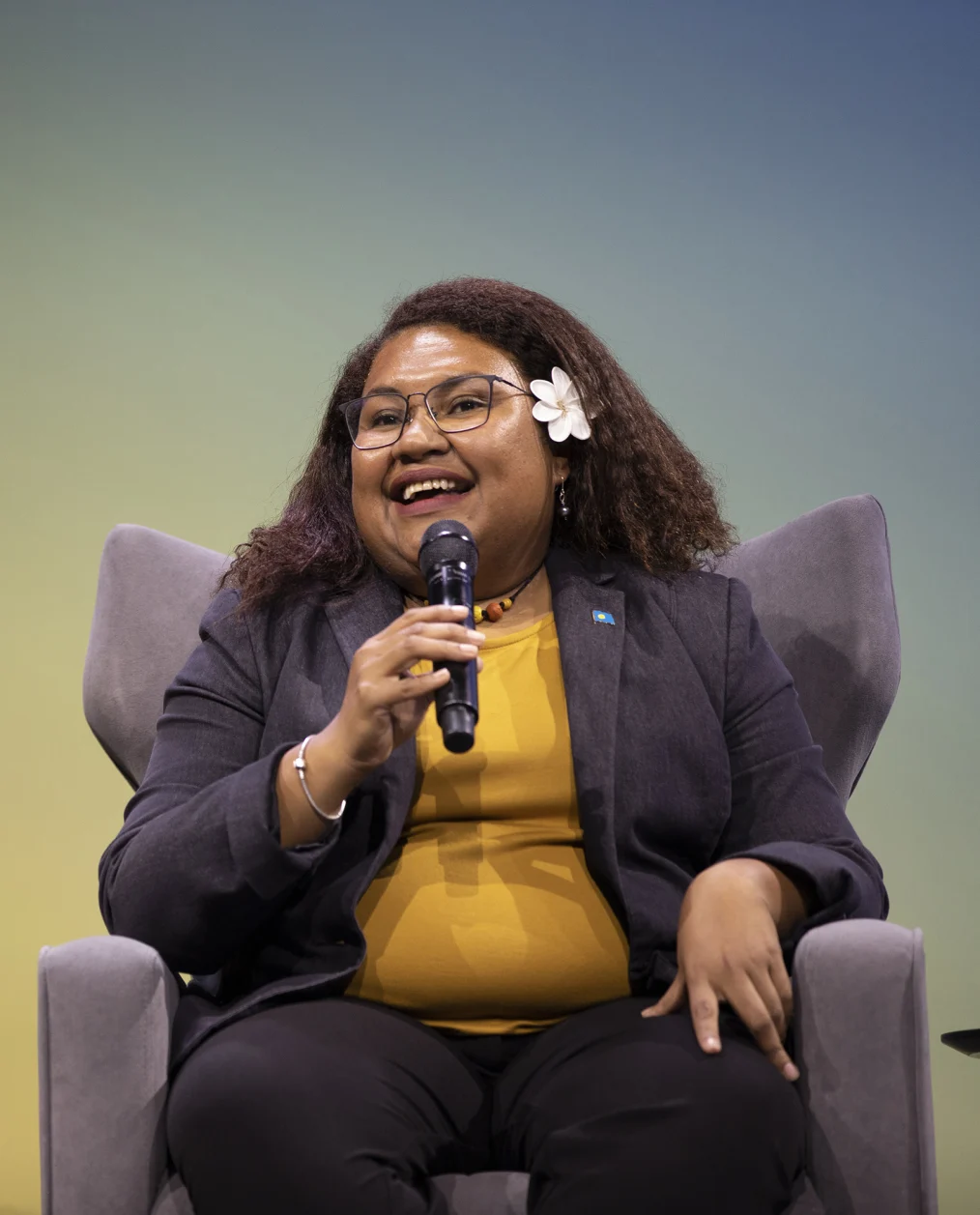 A young woman with a medium-deep skin tone sits and smiles while speaking into a microphone. She wears a yellow top and a gray blazer, glasses and a flower in her hair. In the background, a blue, green and yellow gradient backdrop. 