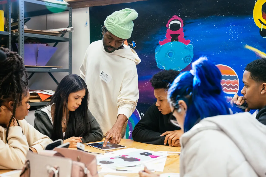 Brandon Breaux, a Black man with a deep skin tone, stands over five seated students. On the table is an iPad and illustrations. He is wearing a mint beanie. Space art is on the wall in the background.