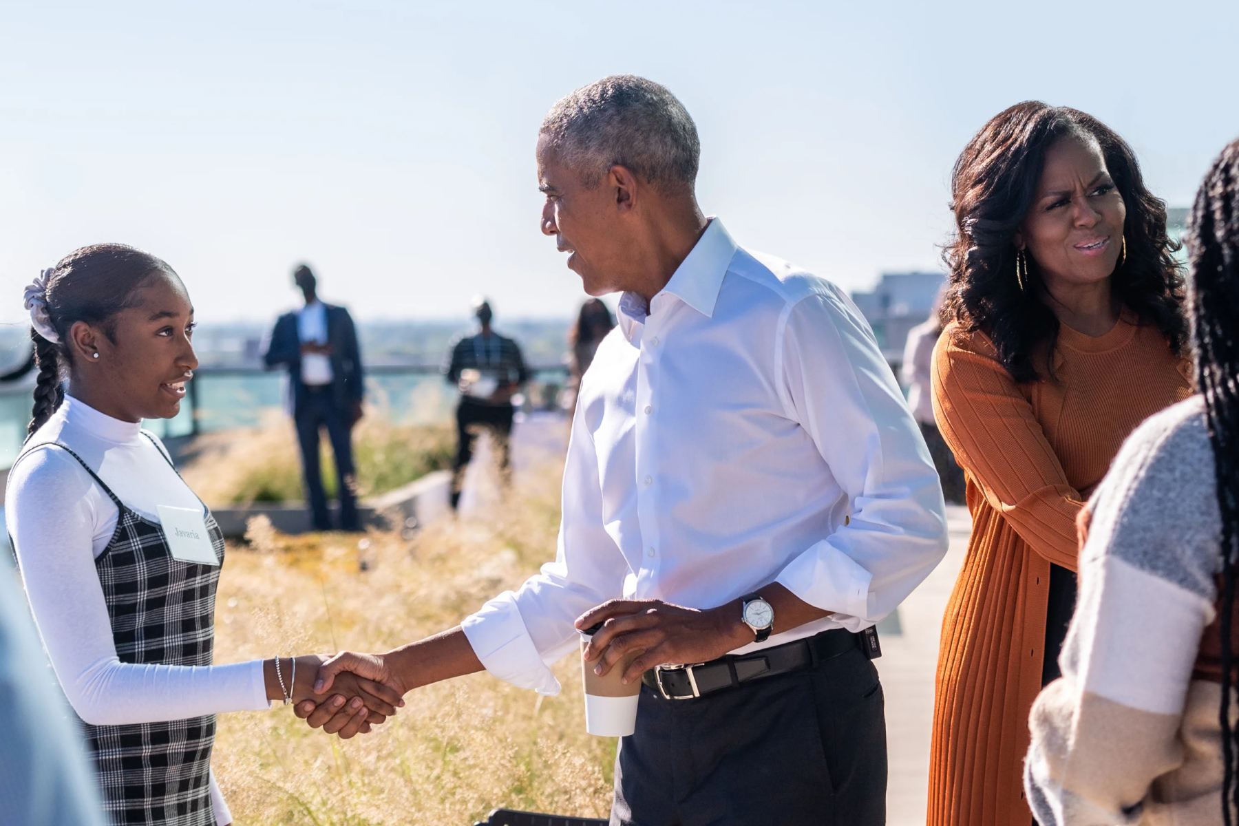 An image of Barack and Michelle Obama shaking the hands of two people while smiling. Barack Obama is shaking the hand of a teenage girl with a deep medium skin tone and dark hair pulled into a braid. Michelle Obama is shaking the hand of a woman with long braids. Barack Obama has a light deep skin tone and gray hair. He is wearing a light colored button down shirt with dark pants and holding a coffee cup in his left hand. Michelle Obama has a medium deep skin tone and curly dark hair. She is wearing an orange sweater and an orange shirt. The picture takes place outside. 