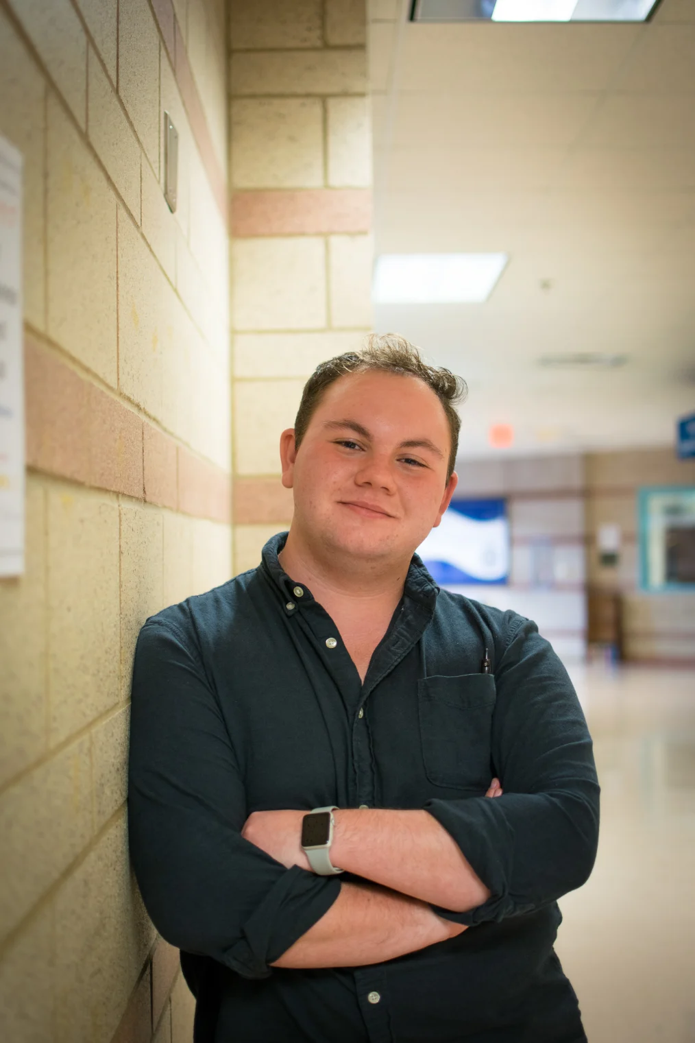 A White man with short and curled light brown hair is leaning against a wall in a building. He is smiling with his mouth closed and his arms folded, showing a watch on his left wrist. He is wearing a dark blue button down shirt with the collar open and the sleeves rolled at the elbow. 