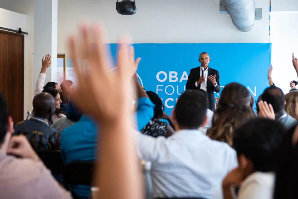 President Barack Obama stands in front of a room of people with hands raised. He holds a microphone. There is a blue sign reading "Obama Foundation" behind him.