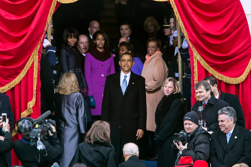 A photo of President Obama in the tunnel on Inauguration Day 2013. He is wearing a black long pea coat. He is surrounded by many people. Behind him is the First Family. In front of him are photographers holing black cameras.