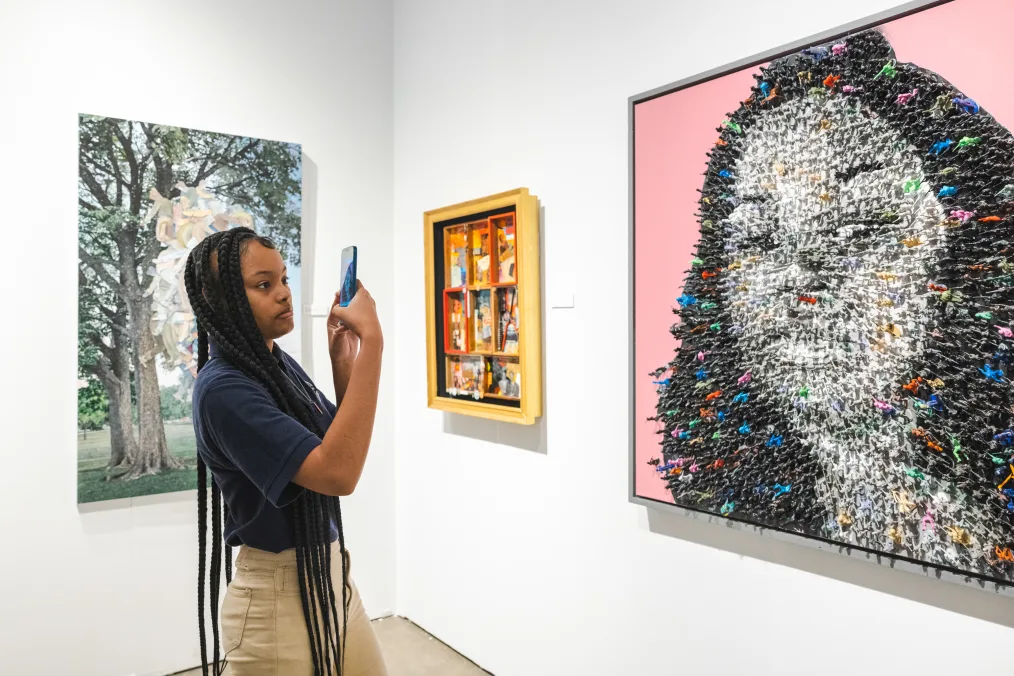 A young woman with a medium deep skin tone stands in front of an art piece tribute to Breonna Taylor. She is taking pictures of the art. Two other pieces are on the wall. The girl has long box braids and is wearing a navy and khaki uniform. 