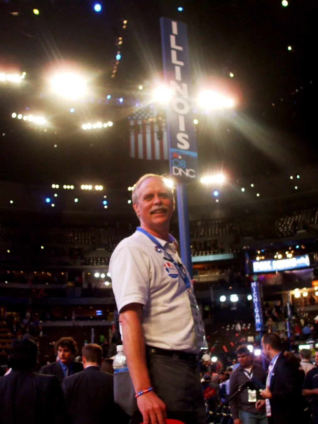 Bright spotlights light up a White man with white hair and white mustache as stands sideways in front of a blue pole with a sign reading "Illinois 08 DNC." People in suit jackets stand behind him. 