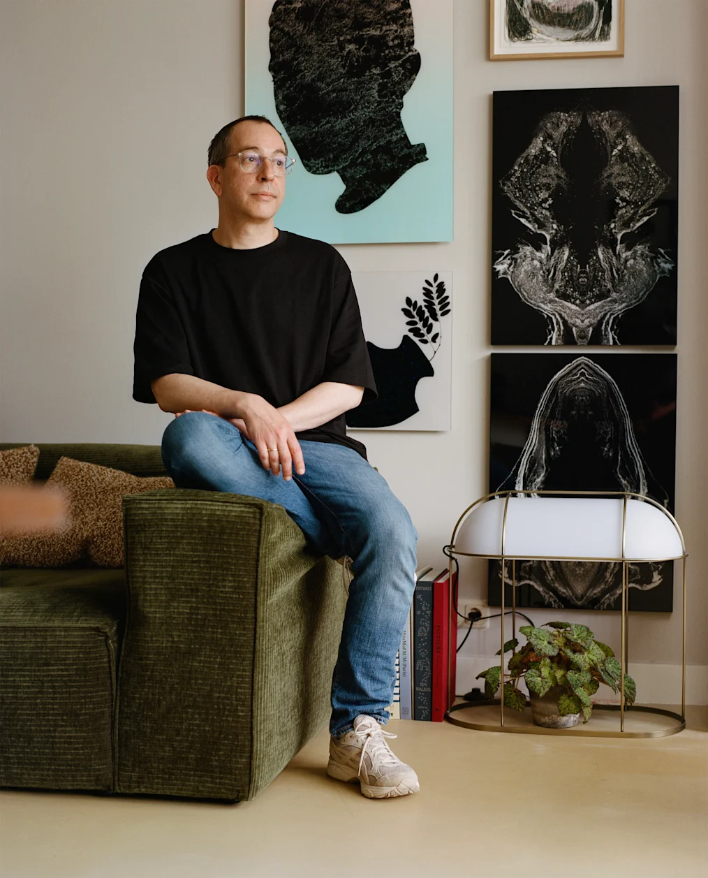 The image is a photographed portrait of Jules Julien. He is sitting with his right leg slightly bent underneath him on the edge of a green corduroy couch with brown pillows. On the wall behind Jules are several art pieces of different sizes and shapes. Sitting on the floor next to Julien is an indoor white plant holder with a light. There is a green plant sitting within the plant holder. Next to the plant holder are three books stacked vertically. Jules has a medium olive complexion and closely cropped dark hair. He is wearing clear rimmed glasses, a black t-shirt, blue denim jeans, and white sneakers.