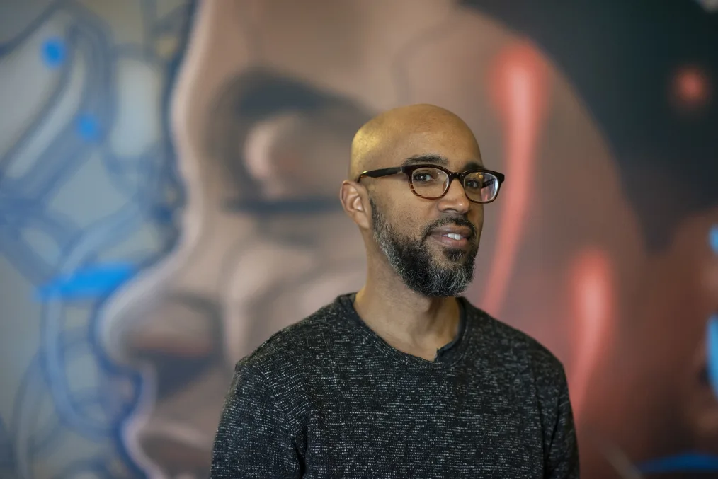 A man who is medium toned, bald, with glasses and a black and grey mixed beard stands infront of a blurred mural. He has a black long sleeve shirt with a pattern of white dashes. His is standing angled slightly to the left and has a gentle smile.