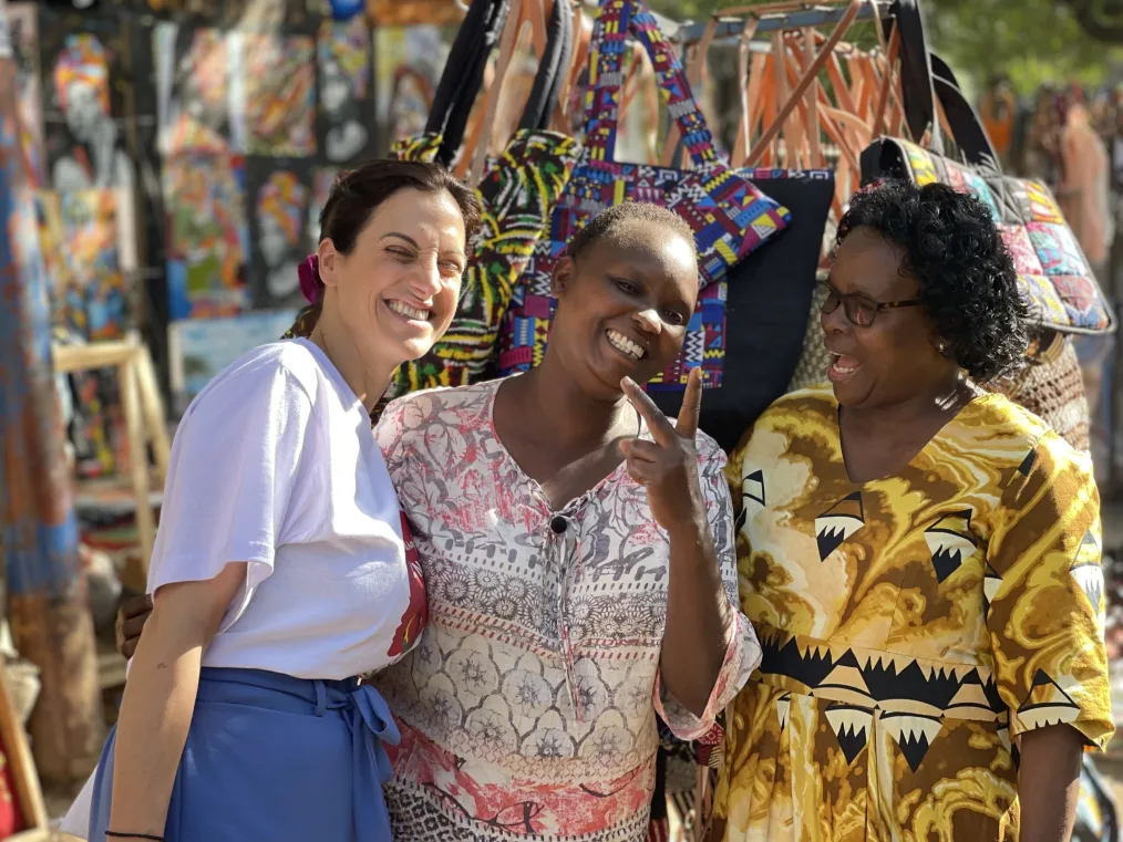 Elpida Kokkota smiles as she stands next to Jame Kalekye Mutambya, a Black woman with a deep skin tone and short hair, and Mercy Musomi, a Black woman with a deep skin tone and short curly hair. Jame holds a peace sign as they stand in front of a booth at a Naroibi cultural market. In the background is art and handmade bags. 