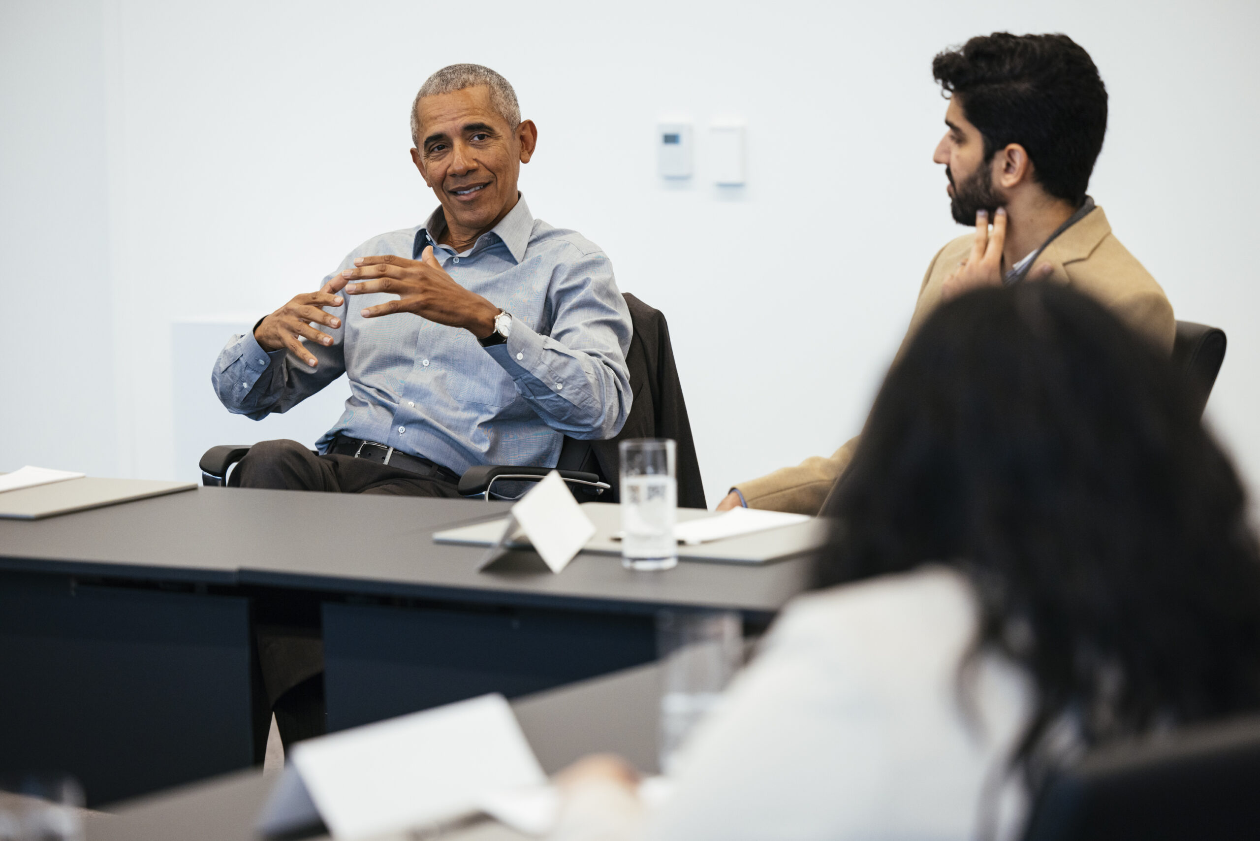 Two Obama Scholars dressed in business casual attire listen attentively President Obama at a table during a roundtable meeting. President Obama gestures with his hands open while speaking.