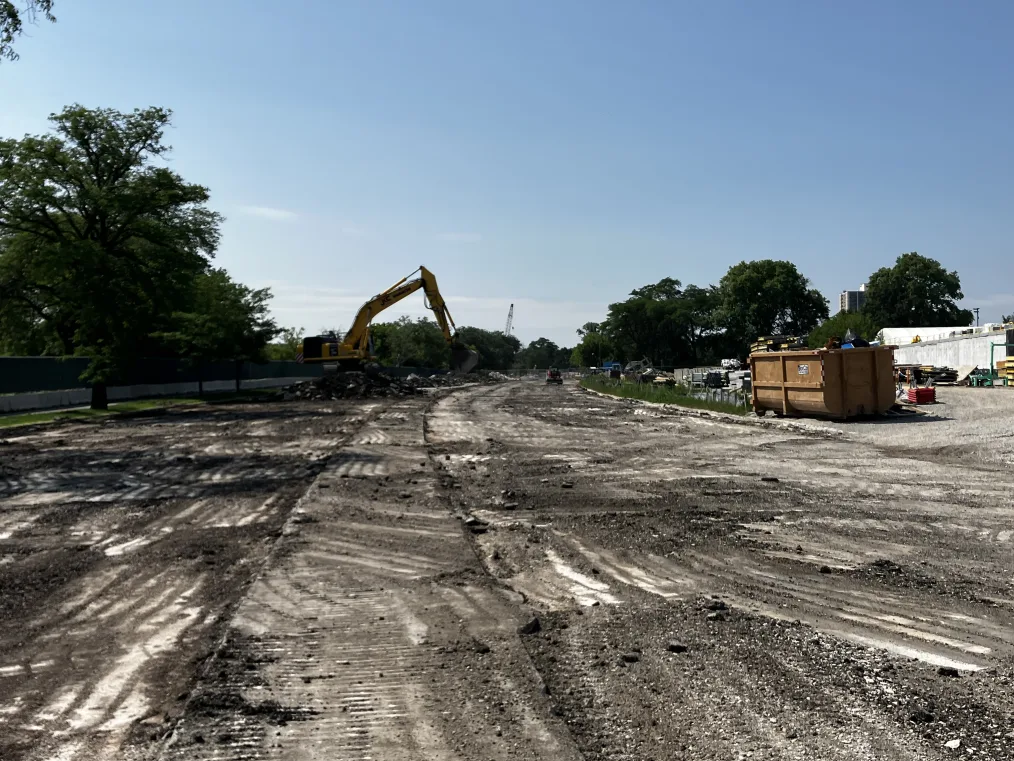 A bulldozer sits in the middle of what was Cornell Drive, where asphalt has been removed revealing a wide dirt pathway. Trees and greenery stand between the former road and the lagoon and on the other side, a rusted metal dumpster filled with debris and various piles of construction materials dot the east side of the main construction zone. 