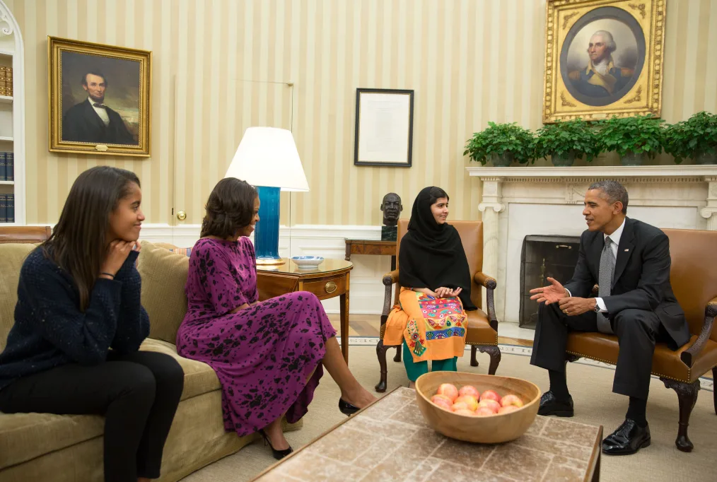 
President Barack Obama, Michelle Obama, Malia Obama, a woman with a deep medium skin tone, and Malala Yousafzai, a woman with a medium skin tone, sit in a room in the White House and have a conversation 