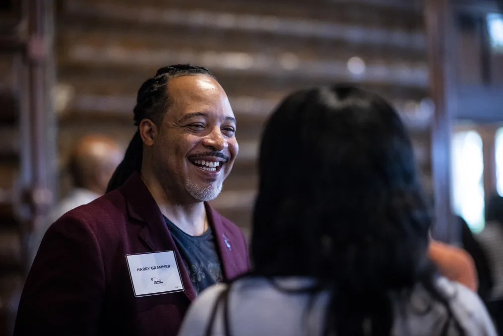 A man with a medium skin tone and a dark red blazer smiles at a person with dark hair.