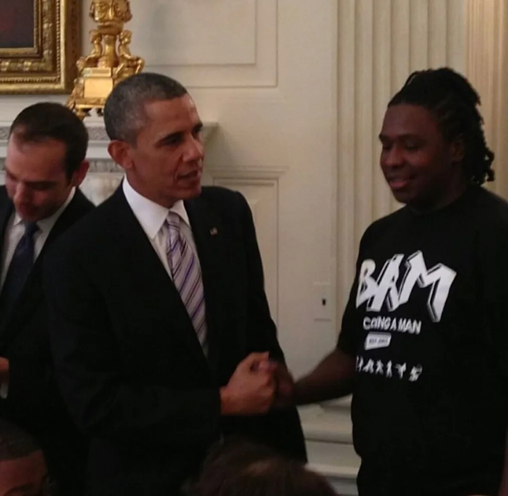  Lazarus Daniels, a young Black man with a dark skin tone and  locs shakes hands with President Obama. President Obama is wearing a dark suit and a striped tie. They are both smiling.