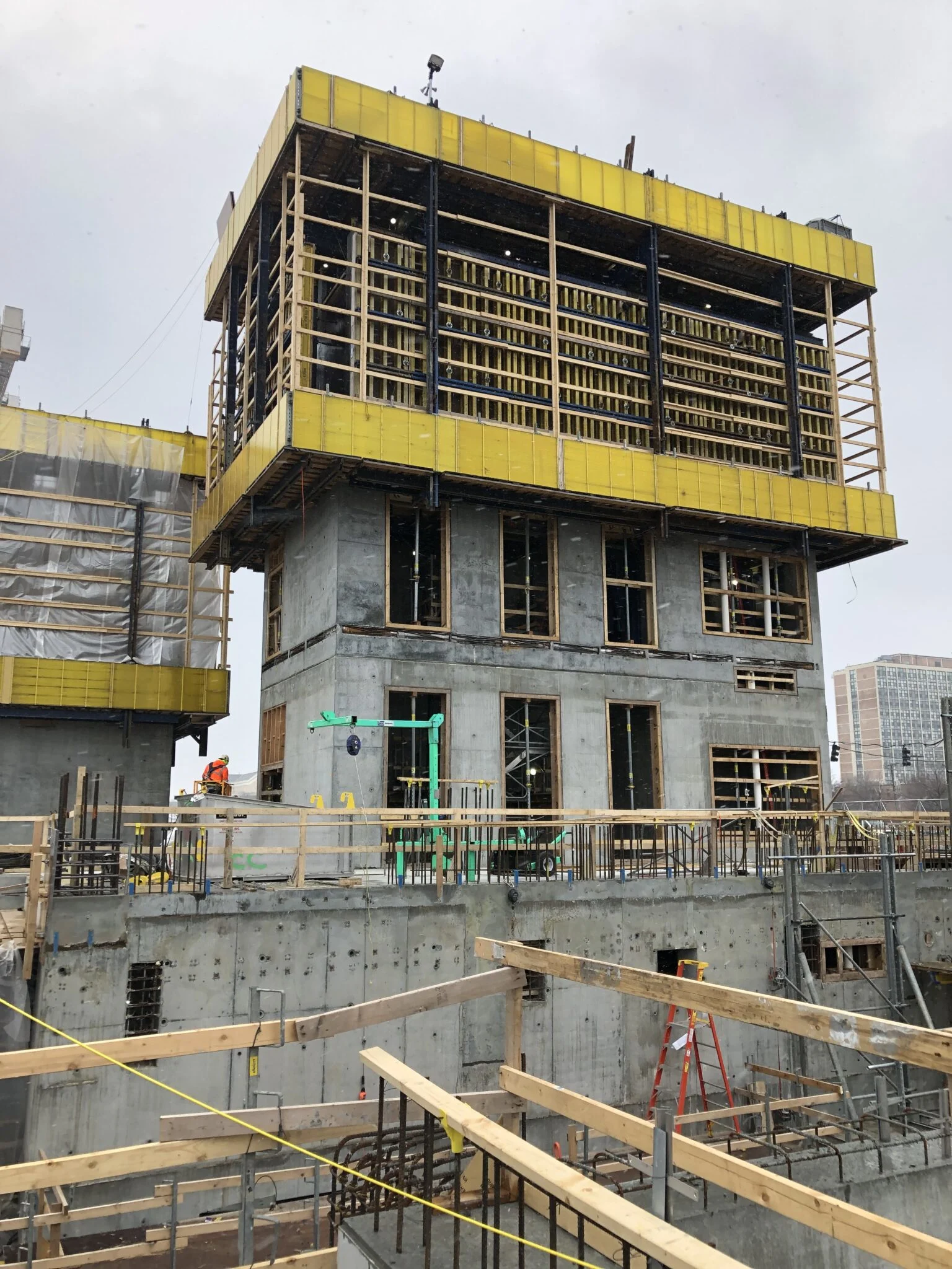 A building being constructed of concrete at the Obama Presidential Center. Beams and rebar can be seen in scaffolding. A worker is wearing orange and working in the background. The sky is gray. 