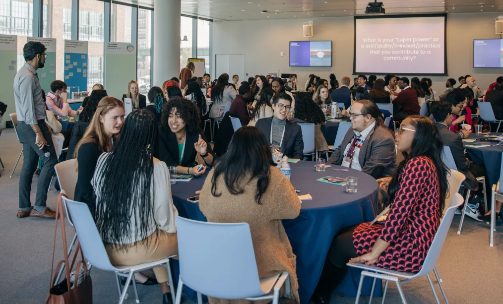 Nick Kennedy sits with six other young adults and fellow Obama-Chesky Voyagers at a round table at the 2022 Democracy Forum in New York City. The young leaders are of all races and the room is full of young people mingling with one another.