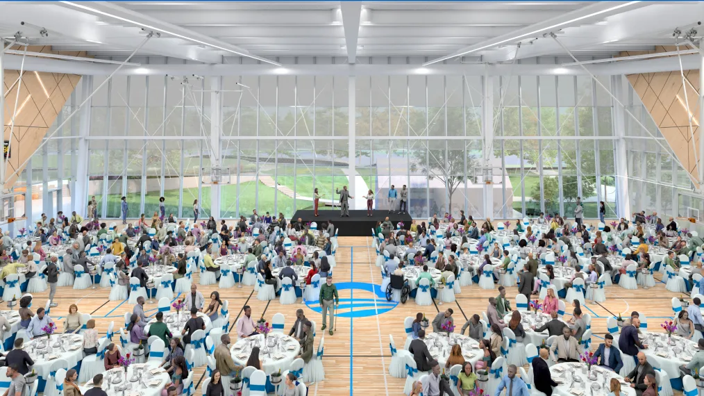 The image is a rendering of the Home Court’s gymnasium. The gymnasium has a wooden floor with basketball court markings and the Obama Foundation logo in blue at center court. The walls of the gymnasium are wood paneling and light colored metal trusses span the ceiling. The wall furthest from the photo is made of rectangular glass windows. Through the windows you can see Jackson Park. On the floor of the gymnasium are dozens of round banquet tables with white tablecloths. Several people are seated at each table. In front of the wall further from the photo is a black stage and podium. Five people stand on the stage. 