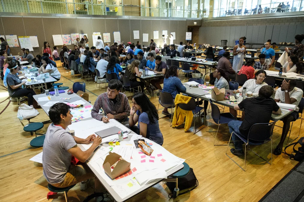 A large group of individuals with a variety of skin tones sit at multiple long gray tables with poster paper and writing utensils. There are brown walls with large white notepad paper and highlighted sticky notes.  