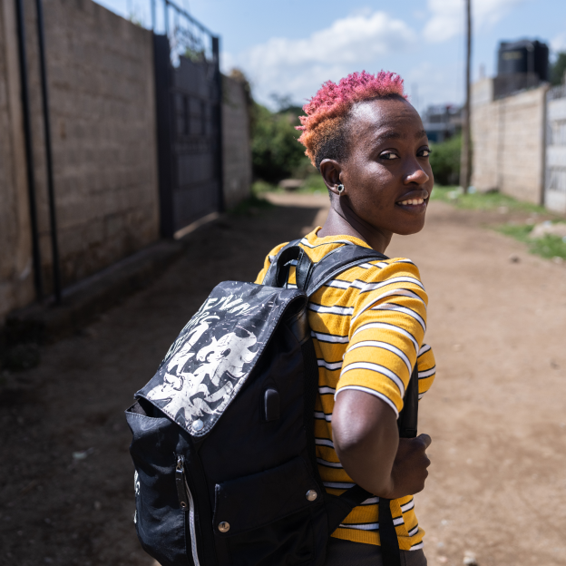 Betty, a young woman with a dark skin tone, turns around and poses for the photo while walking down a dirt path. She is wearing a striped yellow shirt, a black backpack, and has dyed pink hair. 