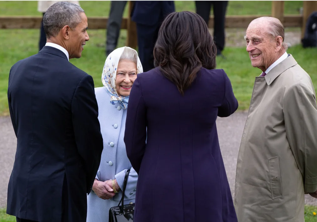 President Obama and Michelle Obama in navy blue business attire smile as they meet the Queen of England, an older woman with a light skin tone with a blue coat, and the King of England, an older man with a light skin tone with a tan coat