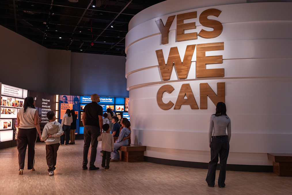 An image of the “Yes We Can” exhibit inside the Museum Building. The exhibit is a large circular structure with words that say “Yes We Can” in wooden letters. An exhibit screen is in the background.  