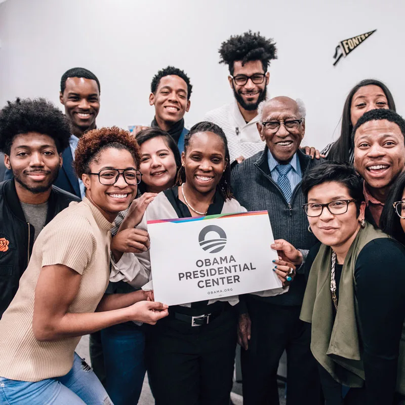 A group of young people smile to camera holding an Obama Presidential Center sign.