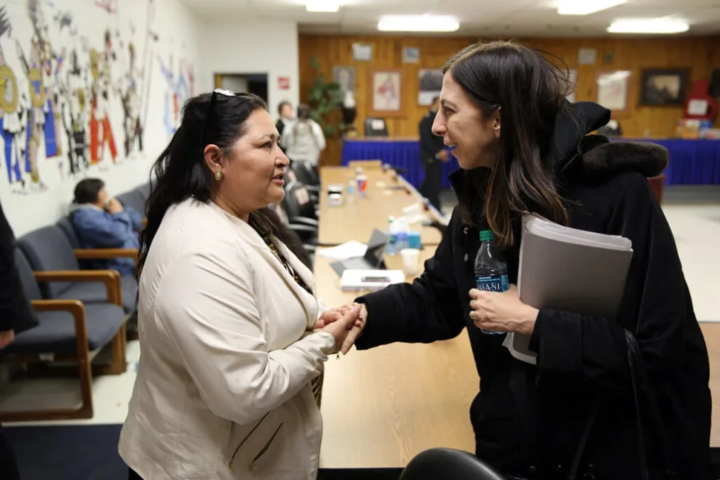Mary Smith, on the right, smiles and shakes hands with a Native woman and tribe member of the Rosebud Sioux Tribe, on the left. The two stand across from each other with a table between them.