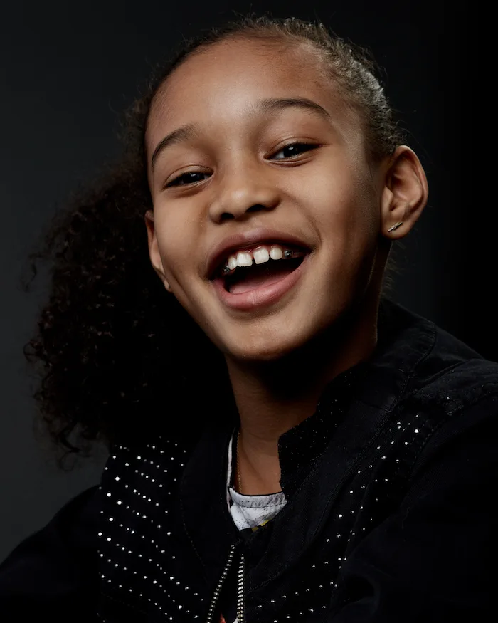 In this portrait, a young black girl with a medium skin tone wearing a black jeweled jacket smiles at the camera 
