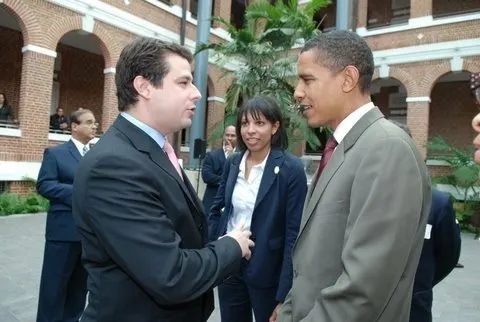 The image is a photo of Myesha Ward, President Barack Obama, and an unidentified man. The unidentified man is speaking to President Obama while Myesha stands in the background. The unidentified man has a pale complexion, short dark hair, and is wearing a dark suit, a white shirt, and a pink tie. President Obama has a light deep complexion, closely cropped black hair and is wearing a grey suit and a red tie. Myesha has a light deep complexion and long dark hair with bangs. She is wearing a dark blue suit and a white shirt underneath.