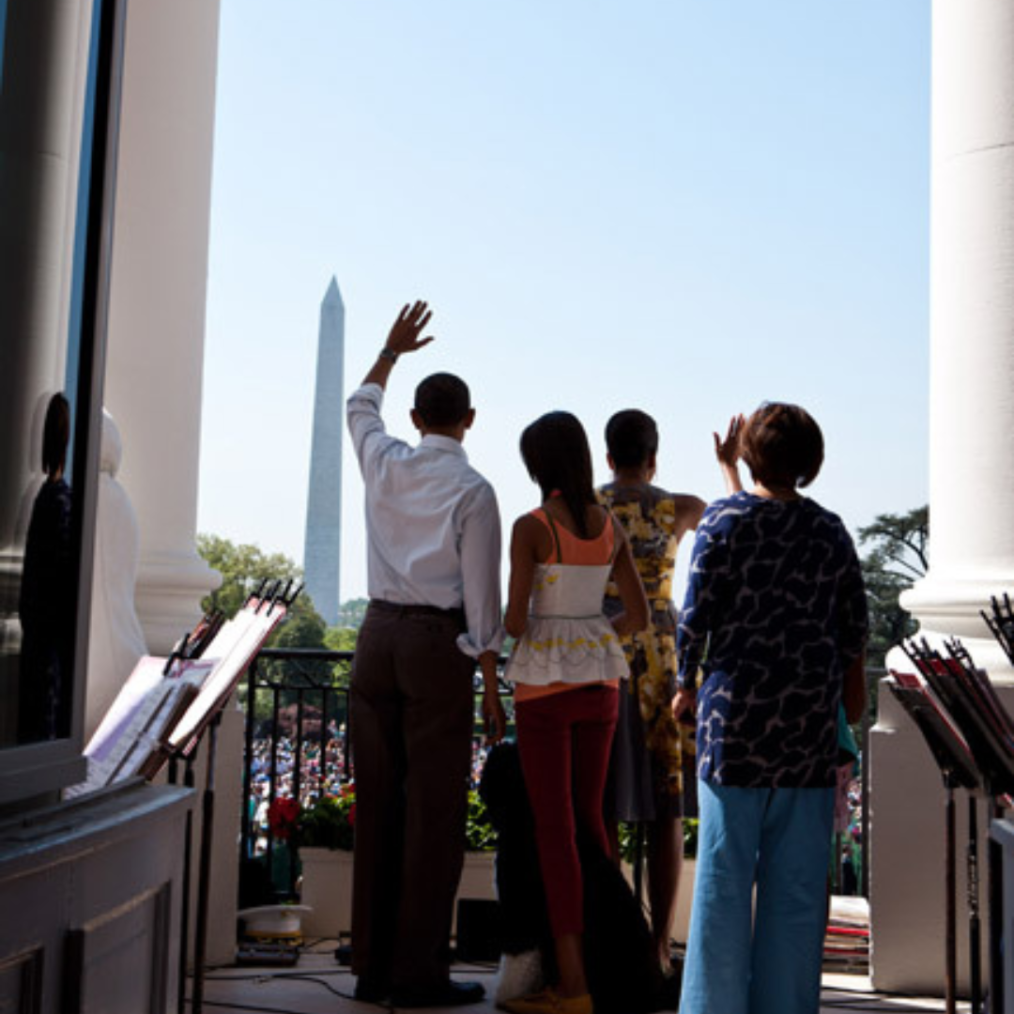 The image is of the back of the Obamas waving to a crowd from the balcony of the White House in the day time. Mrs. Robinson is standing behind the family off to the right side.