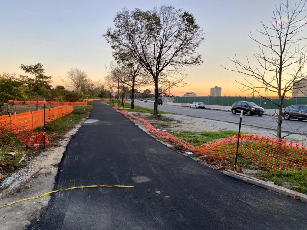 A dark asphalt-paved bike and walking trail is shown in the middle with orange construction fencing on either side of it.