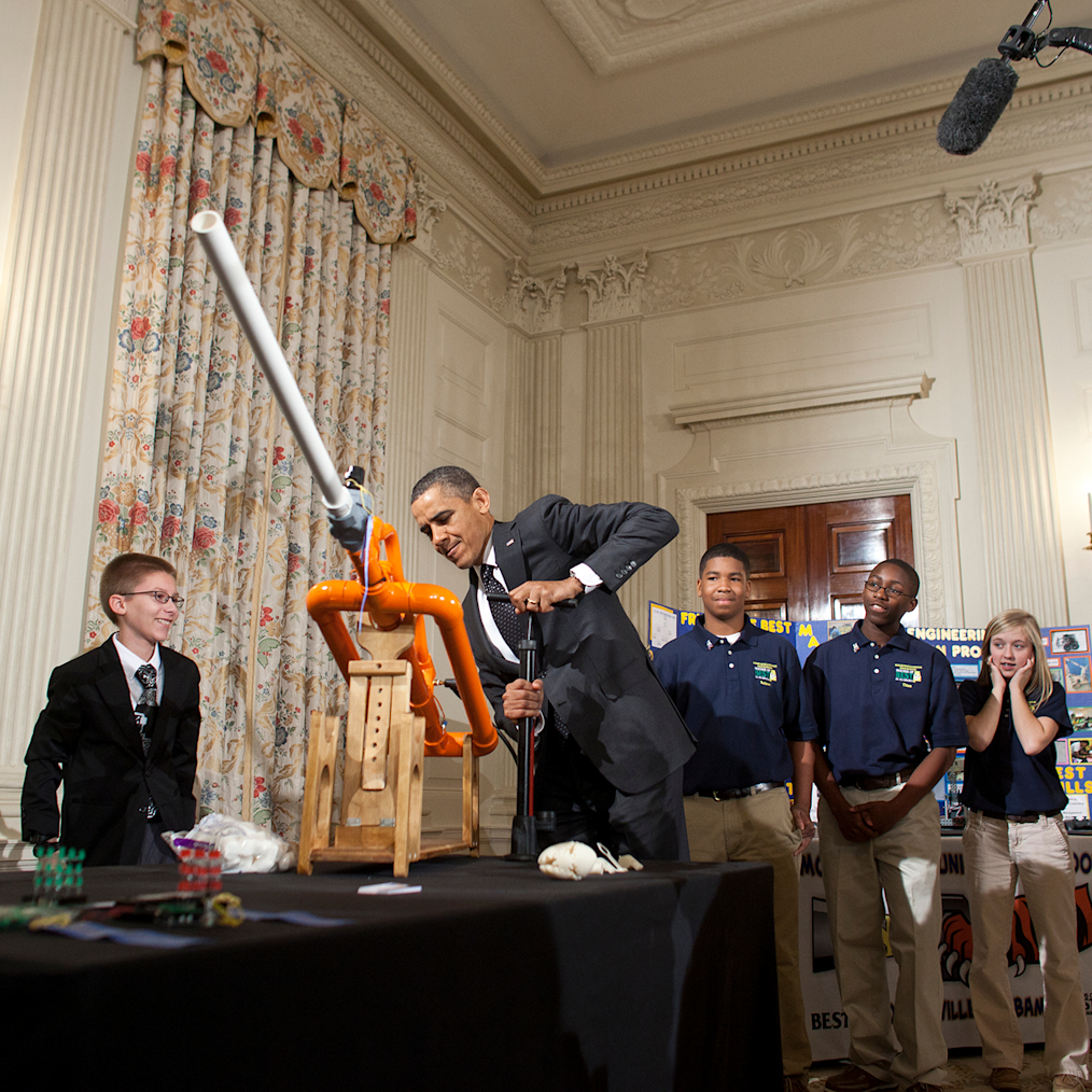 President Barack Obama shooting from a marshmallow cannon at the White House Science Fair. The location is in the White House Dining Room. He is surrounded by several children of different races and ethnicities. 