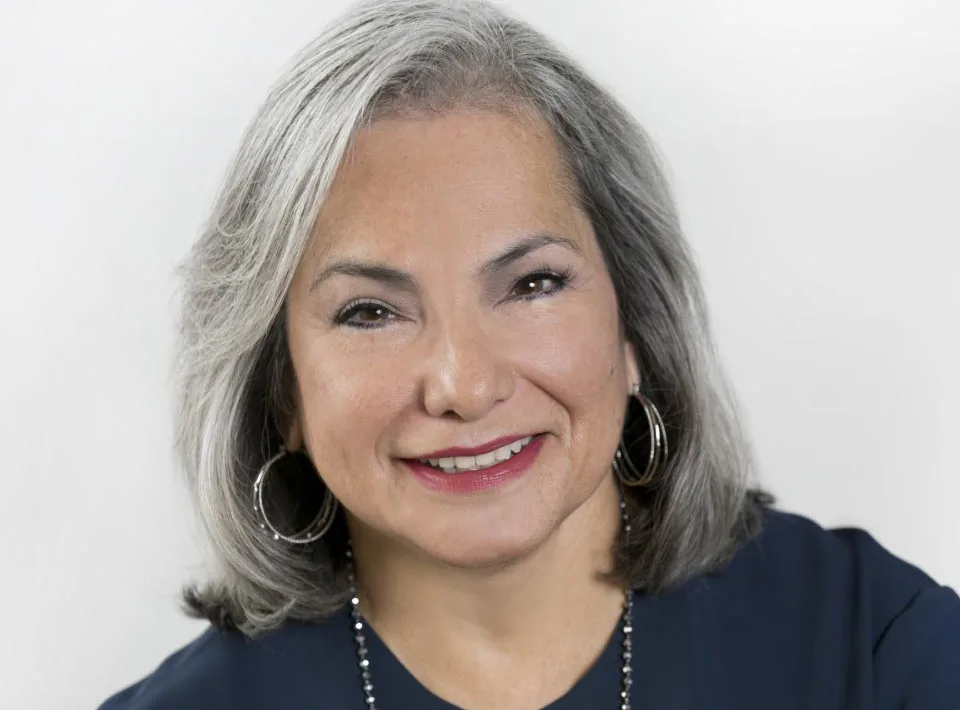 Gloria Castillo, a woman with a light-medium skin tone and short grey hair wearing a dark blue top, smiles toward the camera.