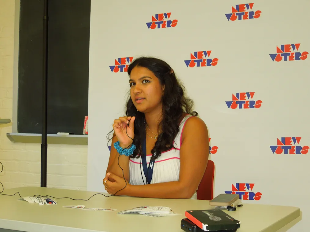 A young adult with medium skin tone and long wavy dark hair sits at a table. They look toward the left with parted lips and raise one hand to their chest. Behind them stands a white banner with a repeated "NEW VOTERS" logo printed in blue and red.
