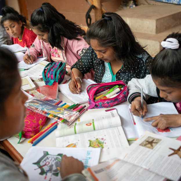 Indigenous girls of the Sacred Valley Project work on school activities together at a long table covered in books, worksheets, colorful markers, and other school supplies.