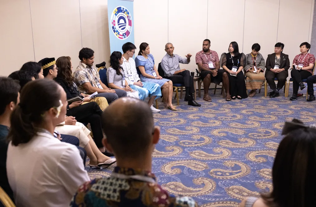 President Obama sits in a circle with people of varying skin tones. He is gesturing with his left hand while speaking. Behind him is a banner that reads "Obama Foundation Leaders Asia-Pacific."