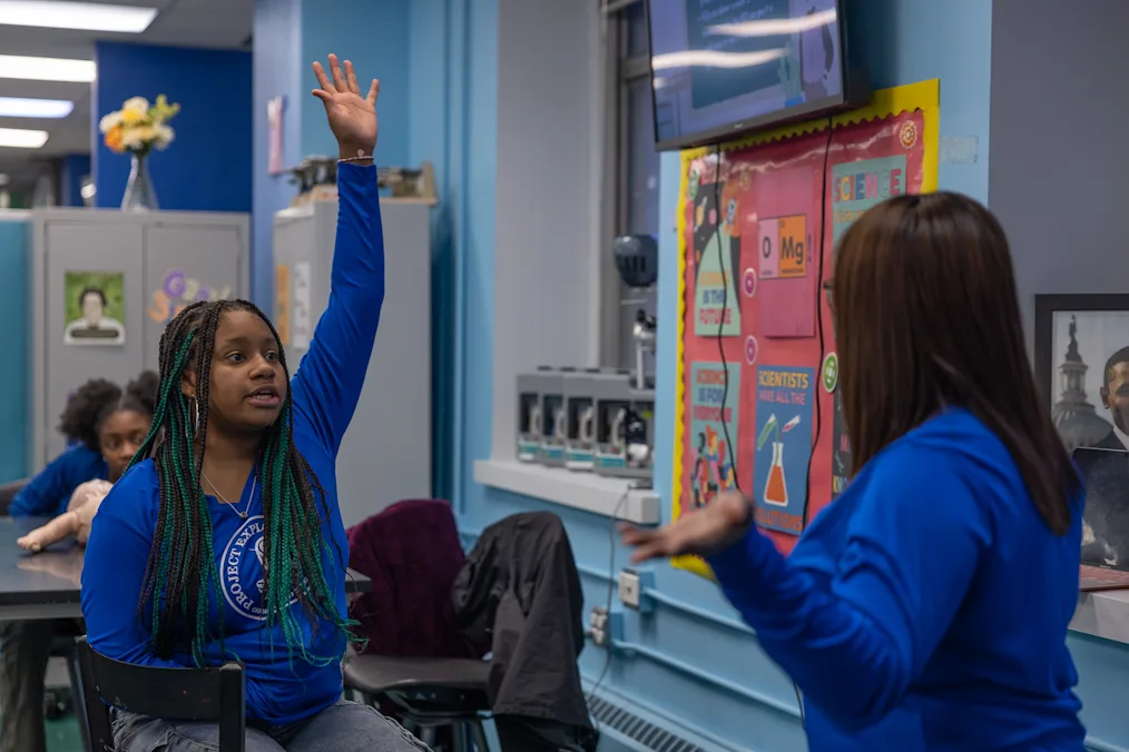 A young Black girl wearing a blue Project Exploration shirt raises her hand in a classroom lab setting as her instructor looks on.