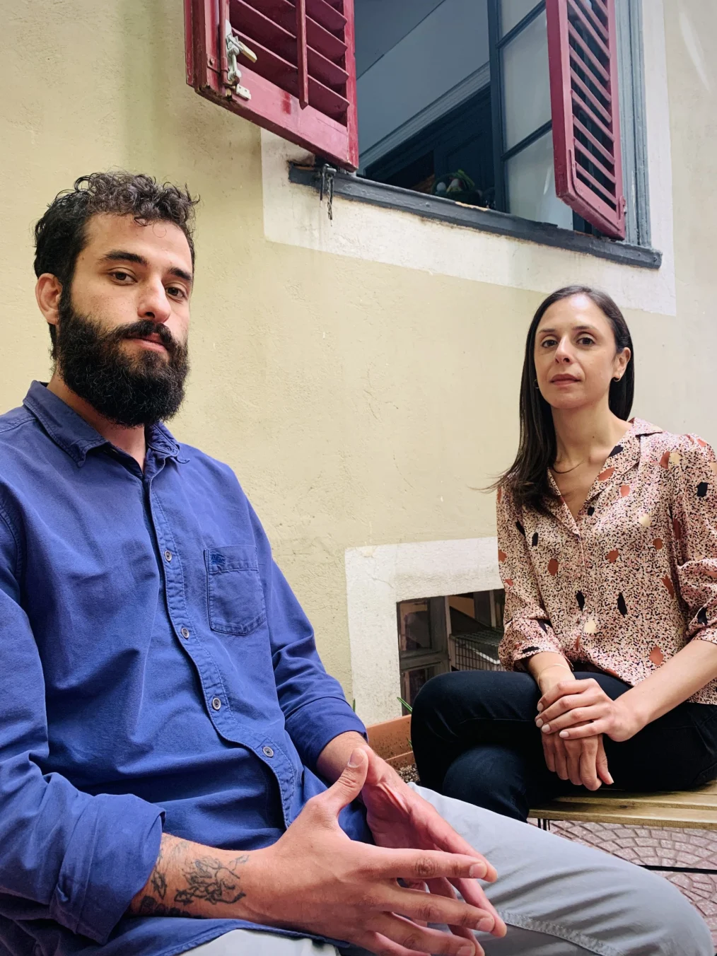 A man and woman with medium-light skin tones sit next to a windowsill posing toward the camera.